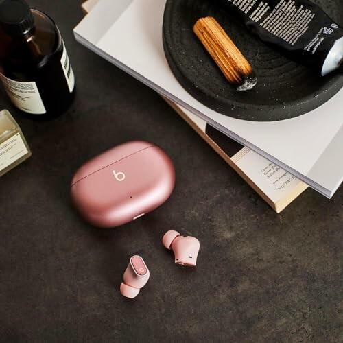 Pink wireless earbuds and case on dark table with books and bottle.